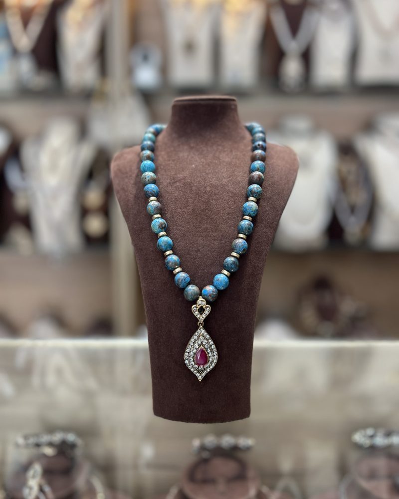 Sautoir en perles texturées bleu turquoise et marron avec rondelles dorées et pendentif goutte serti d'une pierre centrale rouge rubis