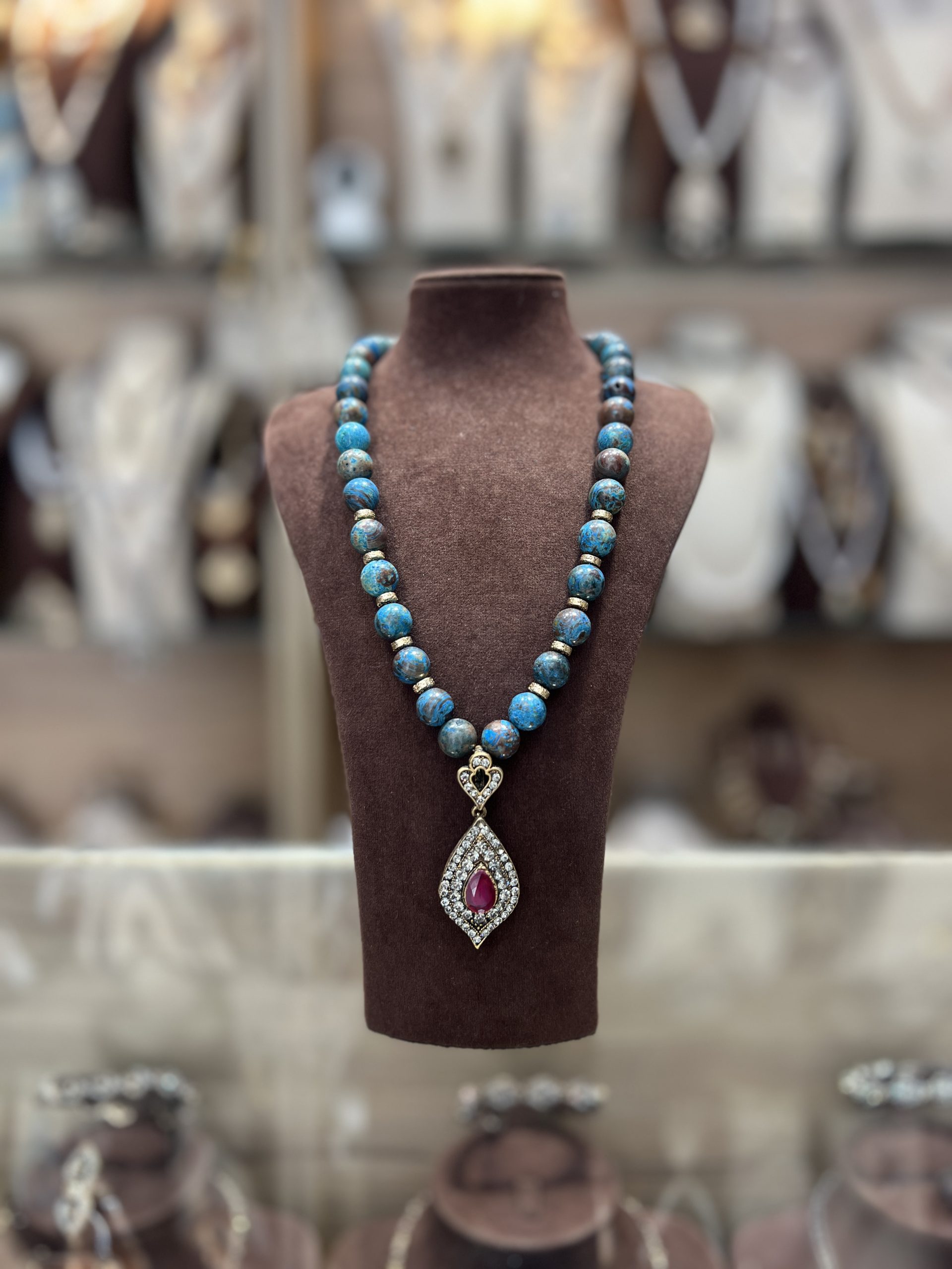 sautoir-perles-bleues-pierre-rubis Sautoir en perles texturées bleu turquoise et marron avec rondelles dorées et pendentif goutte serti d'une pierre centrale rouge rubis
